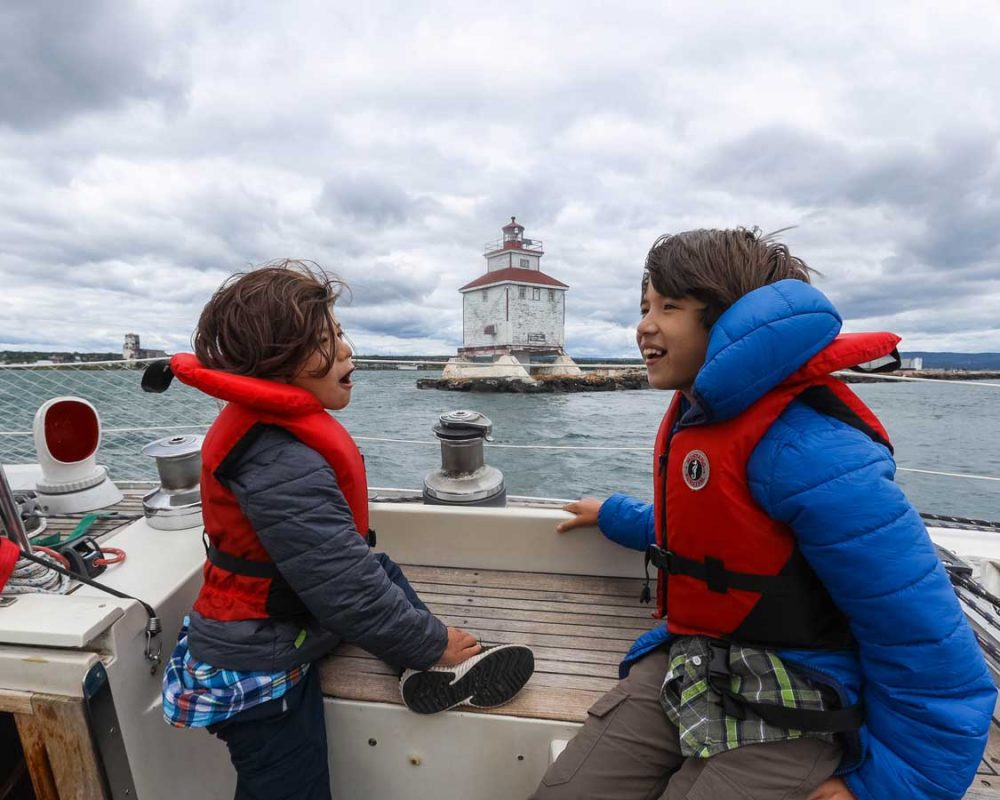 Two boys laugh as they sit on a sailboat boating past a lighthouse in Thunder Bay 