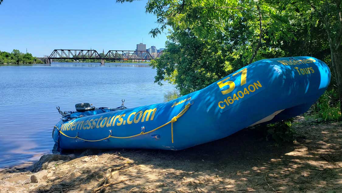 Blue white water raft on a beach with the city of Ottawa in the background