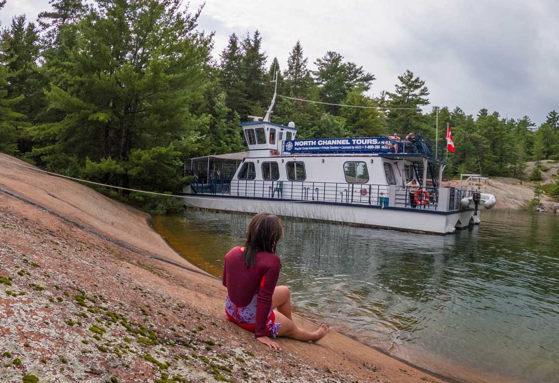 A woman relaxes on the rocks in front of a North Channel Tours boat