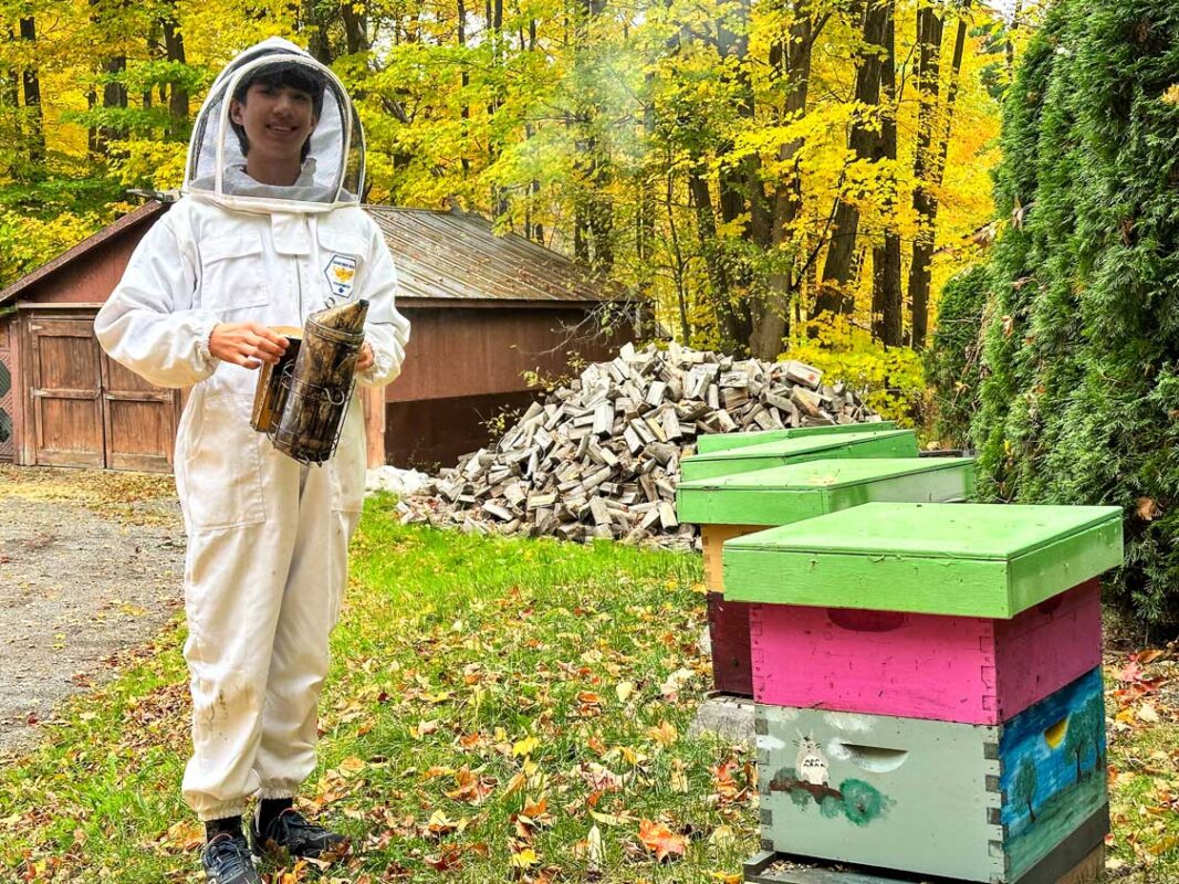 A tall boy smiles while holding a smoker next to a beehive