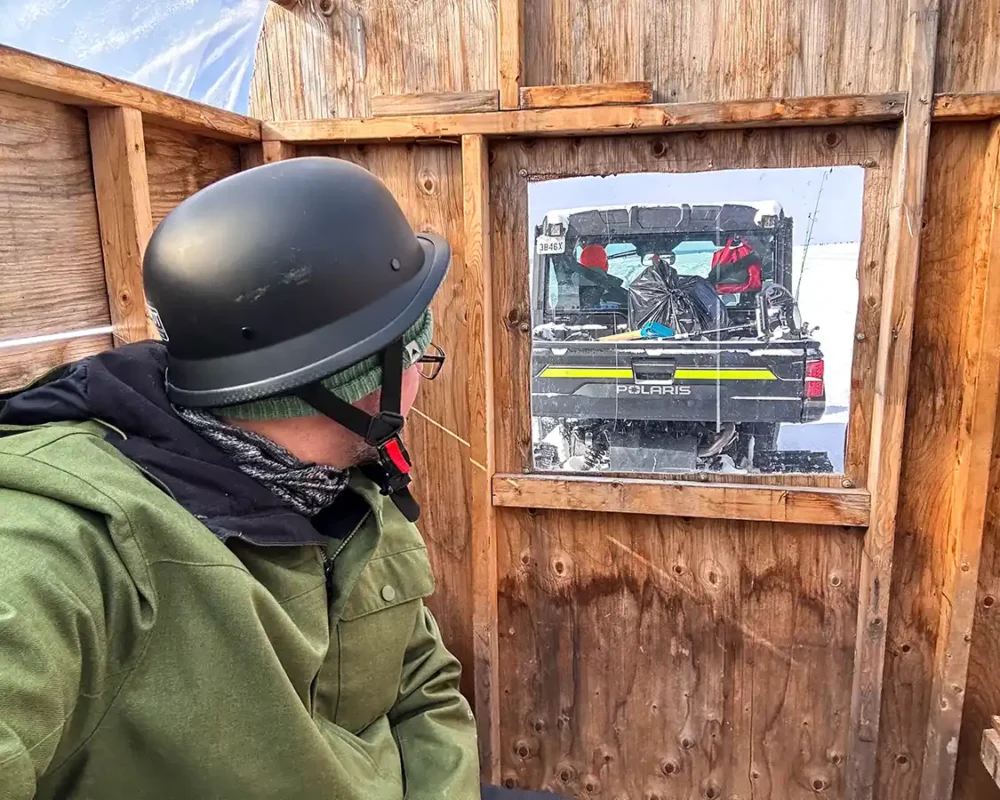 A man wearing a helmet sits in a shed pulled by a side-by-side while ice fishing in North Bay