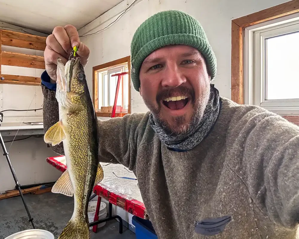 A man in a green toque holds up a small walleye while ice fishing in North Bay